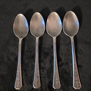 May include: Four silver-toned metal spoons arranged in a row. Each spoon features an oval bowl and a slender handle decorated with a repeating floral pattern and a fleur-de-lis motif at the base. The metal has a matte, slightly tarnished finish.