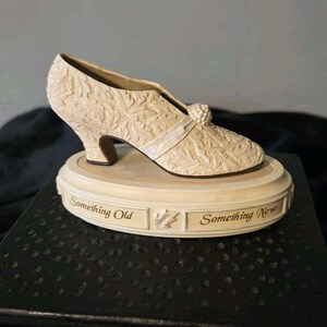 May include: Ivory-colored decorative shoe figurine with intricate floral detailing. The shoe sits atop a round base with the words "Something Old Something New". A ribbon and a decorative embellishment adorn the shoe.