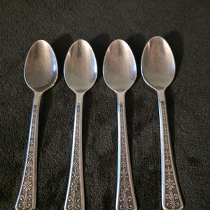 May include: Four silver-toned metal spoons arranged in a row. Each spoon features an oval bowl and a handle decorated with an embossed floral and fleur-de-lis pattern that extends from the base of the bowl to the bottom of the handle.