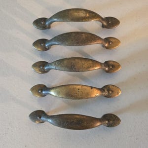 Op de afbeelding: Vijf messingkleurige metalen ladehandgrepen gerangschikt in een verticale kolom. Elke handgreep heeft een gebogen, langwerpig middengedeelte met een afgerond, druppelvormig bevestigingspunt aan beide uiteinden, met een verweerde patina-afwerking.