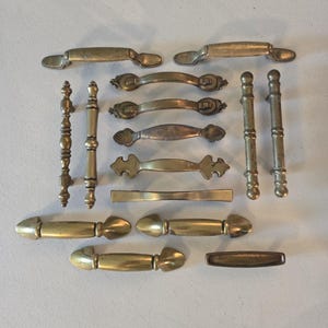 Op de afbeelding: Een verzameling van twaalf messing kastgrepen en handgrepen in verschillende vormen en maten. Het assortiment bevat rechte stangen, gebogen grepen met decoratieve uiteinden en cilindrische staven met textuurdetails, allemaal met een metalen afwerking.