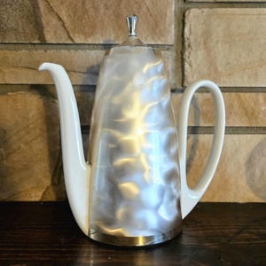 Pode incluir: Uma cafeteira prateada vintage com uma alça e bico brancos. A cafeteira tem um acabamento em metal escovado e um pequeno botão prateado na tampa. A cafeteira está sobre uma superfície de madeira.