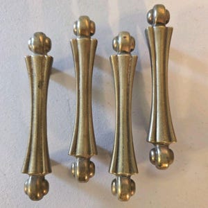 May include: Four vertical brass cabinet pulls arranged in a row. Each pull features a central elongated body with a fluted design, capped at both the top and bottom with a rounded, spherical finial. The metal has a muted, aged gold finish.