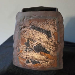 May include: A dark brown ceramic vase with a rectangular, flattened shape. The front surface features a textured, relief-style design depicting two fish swimming among aquatic plants, rendered in shades of brown, tan, and black clay.