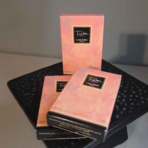 Puede incluir: Tres cajas rosas de perfume Trésor de Lancôme apiladas sobre una superficie negra. Cada caja tiene una etiqueta negra con ribete dorado y el nombre "Trésor" y "Lancôme Paris" en texto blanco. Las cajas tienen un diseño floral rosa.