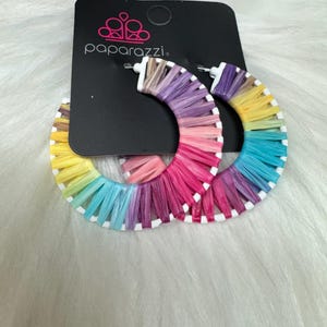 May include: A pair of rainbow-colored hoop earrings. The earrings are made of white hoops wrapped with colorful threads in shades of yellow, green, blue, pink, and purple. The earrings are attached to a black tag with the word "paparazzi" printed on it.