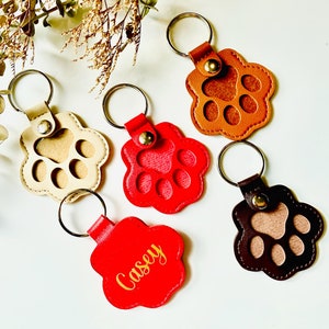 May include: Five leather paw print keychains with metal rings. One keychain is red with the name "Casey" in gold lettering. Another keychain is brown with a tan paw print. Two keychains are tan with brown paw prints. One keychain is brown with a tan paw print.