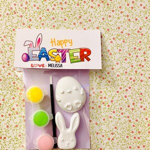 May include: A Happy Easter craft kit with a white ceramic bunny and egg, paintbrushes, and three paint pots in yellow, green, and pink. The kit comes with a card that says "Happy Easter" and "Love: Melissa".
