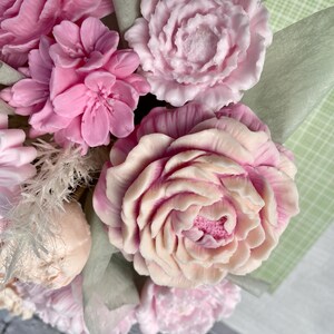 Puede incluir: Un ramo de flores de jab&oacute;n rosas y blancas dispuestas en un envoltorio de papel verde y blanco. Las flores est&aacute;n hechas para parecer peon&iacute;as y rosas.
