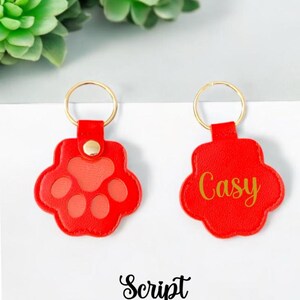 May include: Two red leather keychains with gold rings. One keychain has a paw print design and the other has the name "Casey" written in gold script.