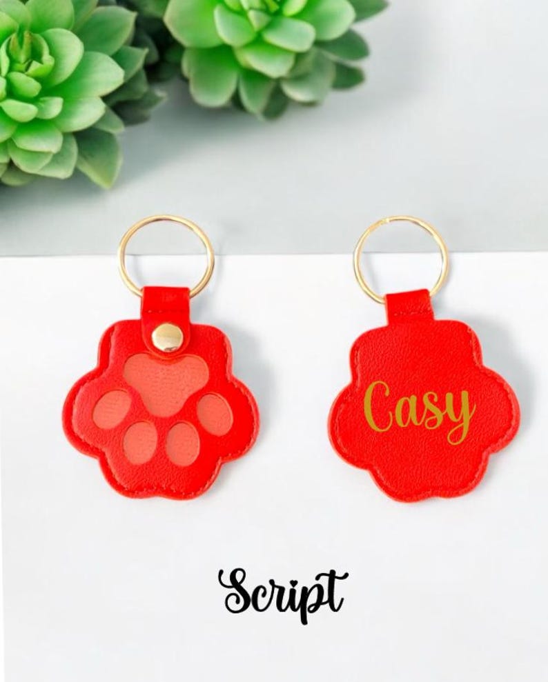 May include: Two red leather keychains with gold rings. One keychain has a paw print design and the other has the name "Casey" written in gold script.