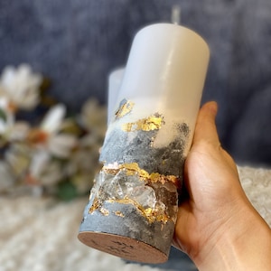 May include: A white pillar candle with a gray and gold decorative layer. The candle has a textured surface and is decorated with gold leaf and clear crystals.