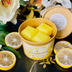 May include: A brown paper cup filled with three yellow lemon scrub cubes. The cup has a label that reads "Lemon Scrub Cubes" and "All Natural Ingredients".
