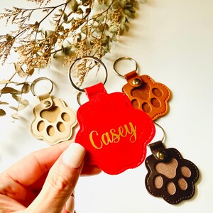 May include: Three leather paw print keychains with gold rings. One keychain is red with the name "Casey" in gold lettering. One keychain is brown with a brown paw print. One keychain is beige with a brown paw print.