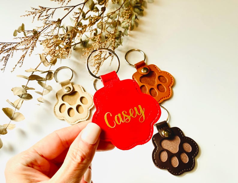 May include: Three leather paw print keychains with gold rings. One keychain is red with the name "Casey" in gold lettering. One keychain is brown with a brown paw print. One keychain is beige with a brown paw print.