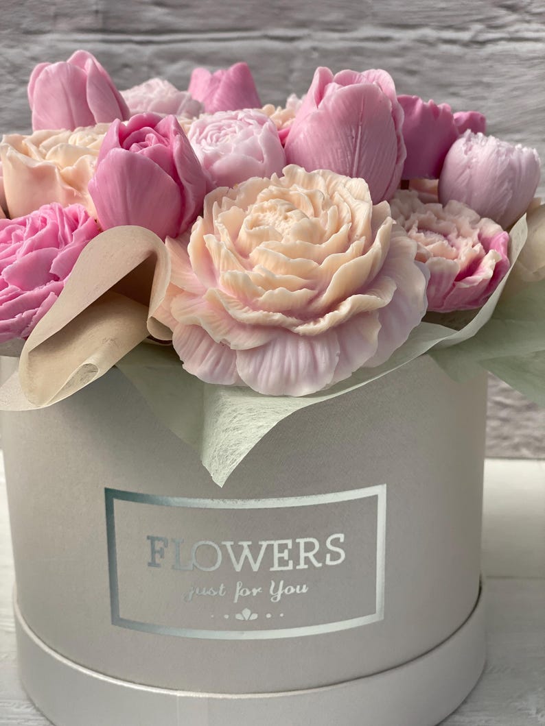 Puede incluir: Un ramo de flores de jab&oacute;n rosas y blancas dispuestas en una caja de regalo redonda blanca con una etiqueta plateada que dice "FLOWERS just for you".