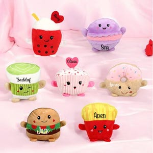 May include: A collection of plush food toys, including a red bubble tea, a purple macaron, a green matcha latte, a pink cupcake, a tan donut, a burger, and a red french fry container. Each toy has a smiling face and a name tag.