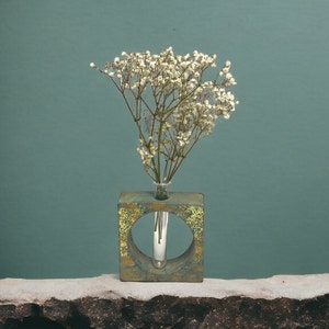 May include: A square, weathered-look vase with a circular opening, holding a small glass vial with white flowers. The vase has a green and gold patina, set against a teal background.