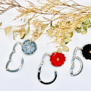 May include: Three silver handbag hooks with different colored fabric flower accents. The hooks are in the shape of a 'C' and have a spring mechanism. The flowers are red, blue, and black.