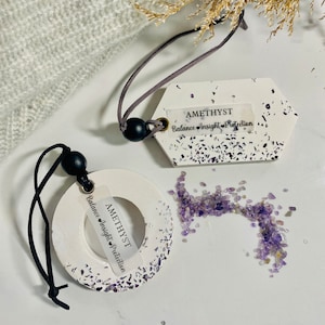 May include: Two white ceramic car air fresheners with purple glitter and the words "Amethyst Balance Insight Protection" on them. One is shaped like a circle and the other is shaped like a tag. There are loose purple crystals scattered around the air fresheners.