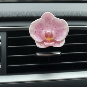 May include: A pink orchid car air vent clip. The flower has a detailed design with a yellow center and pink spots. The clip is attached to a black car vent.