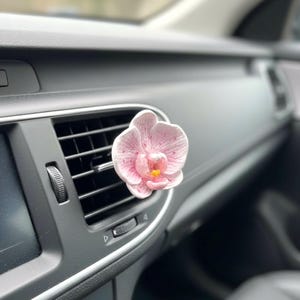 May include: A pink and white orchid-shaped car air freshener is clipped to a car vent. The flower has a yellow center and is set against a dark gray dashboard. The car interior is visible in the background.