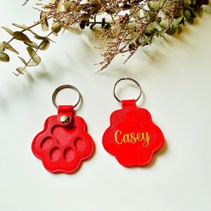 May include: Two red leather keychains shaped like paw prints. One keychain has the name "Casey" written in gold lettering. The other keychain has a design of five circles inside the paw print.