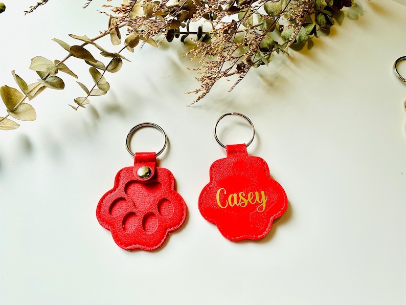 May include: Two red leather keychains shaped like paw prints. One keychain has the name "Casey" written in gold lettering. The other keychain has a design of five circles inside the paw print.