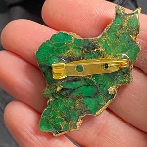 May include: A green and gold resin brooch shaped like the outline of a country. The brooch has a gold pin on the back.