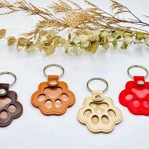 May include: Four leather paw print keychains in different colors: brown, orange, beige, and red. Each keychain has a metal ring and a clear window for a photo.