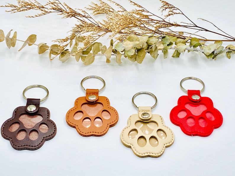 May include: Four leather paw print keychains in different colors: brown, orange, beige, and red. Each keychain has a metal ring and a clear window for a photo.