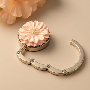May include: A gold-toned handbag hook with a delicate peach-colored flower design. The hook is foldable and has a spring-loaded mechanism.