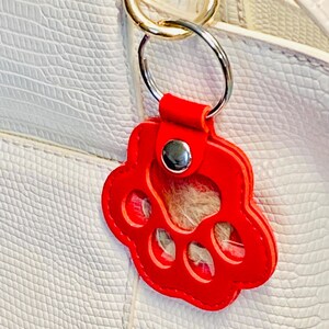 May include: Red leather paw print keychain with a clear plastic insert. The keychain is attached to a gold-colored metal ring.