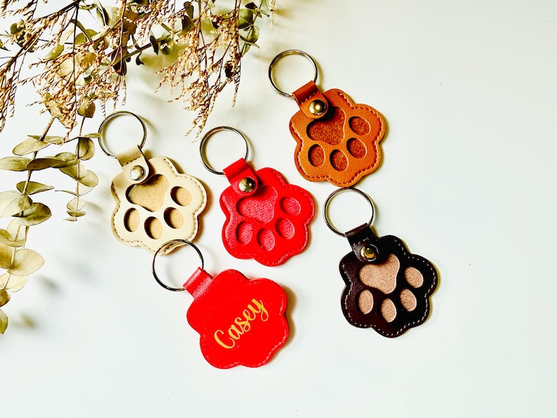 May include: Five leather paw print keychains with metal rings. One keychain is red with the name "Casey" in gold lettering. Another keychain is brown with a tan paw print. Two keychains are tan with brown paw prints. One keychain is brown with a tan paw print.
