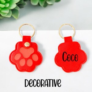 May include: Two red leather keychains with gold key rings. One keychain has a paw print design and the other has the name "Coco" written on it. The keychains are labeled "DECORATIVE".