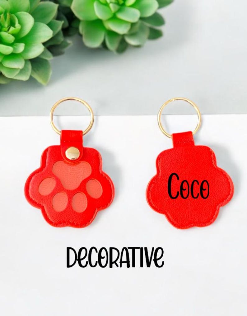 May include: Two red leather keychains with gold key rings. One keychain has a paw print design and the other has the name "Coco" written on it. The keychains are labeled "DECORATIVE".