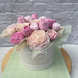 Puede incluir: Un ramo de flores de jab&oacute;n rosa y blanco dispuestas en una caja blanca con la palabra "FLOWERS" impresa en la parte delantera. La caja est&aacute; sobre una superficie a cuadros verde y blanco.