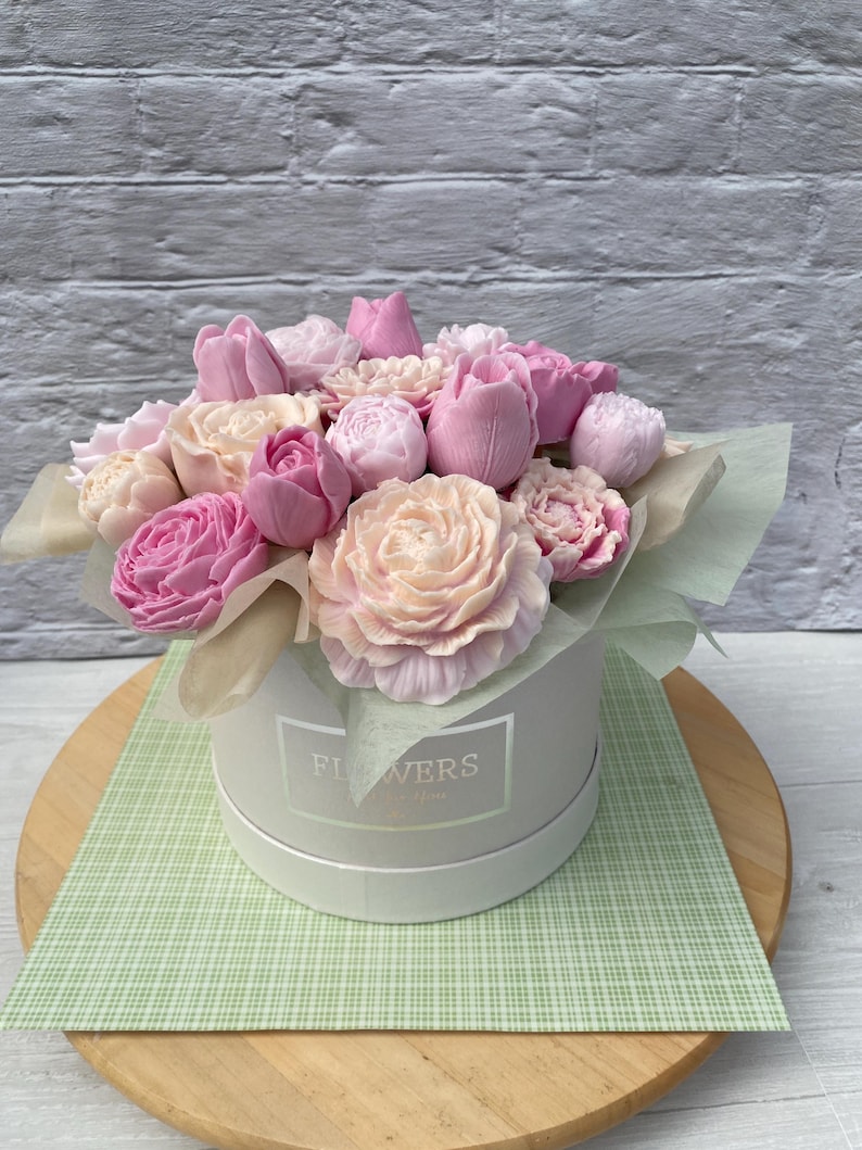Puede incluir: Un ramo de flores de jab&oacute;n rosa y blanco dispuestas en una caja blanca con la palabra "FLOWERS" impresa en la parte delantera. La caja est&aacute; sobre una superficie a cuadros verde y blanco.