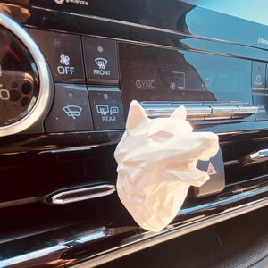 May include: White ceramic wolf head car air freshener. The wolf is facing right and has an open mouth. The air freshener is sitting in the air vent of a car.