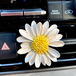 May include: White daisy car air freshener with a yellow center. The daisy is attached to a black car vent.