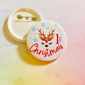 May include: White button with a red reindeer with closed eyes and antlers, snowflakes, and the text "1st Christmas" in red.