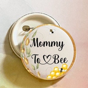 May include: A white button with a gold border and a bee design. The text "Mommy To ♥ Bee" is printed in black.