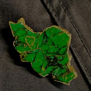 May include: A green and gold pin shaped like the outline of a country with a small heart in the center.