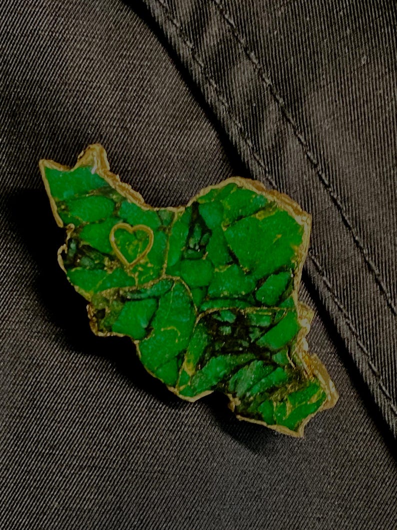 May include: A green and gold pin shaped like the outline of a country with a small heart in the center.