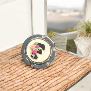 May include: A silver-toned metal purse hook with a white background and a black letter F with pink flowers. The hook is on a woven surface.