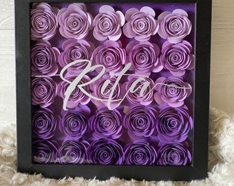 May include: A black framed shadow box with a glass front filled with purple paper flowers. The name "Rita" is written in white cursive lettering in the center of the box.