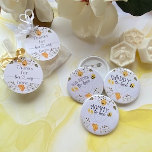 May include: Four round white and yellow paper favor tags with bee and honeycomb designs. The tags say "Thanks for beeing here" and "Big Sister to bee", "Mommy to bee", and "Daddy to bee".