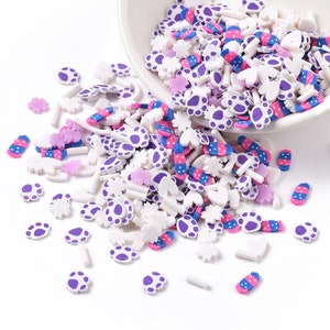 May include: A selection of colourful and white plastic sprinkles, including paw prints, hearts, flowers, and stripes. The sprinkles are scattered on a white surface.