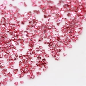 May include: Pink iridescent glass glitter, scattered on a white surface. The glitter is made of small, irregular pieces of glass.
