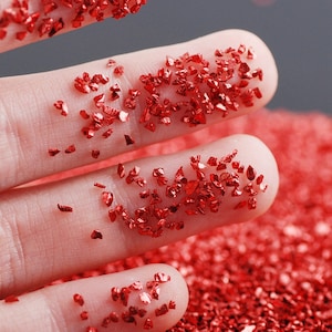May include: Close-up of red crushed glass pieces on a person's fingers. The small, irregularly shaped pieces are scattered across the skin, with a larger pile of the same material in the background. The glass has a reflective, shiny surface.